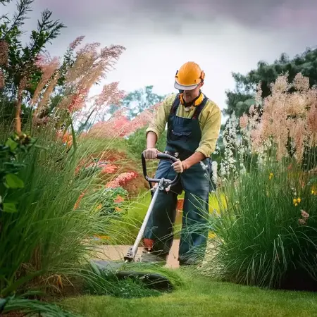 Gärtner mit Schutzhelm trimmt Rasen im gepflegten Garten mit Motorsense und Blumen.